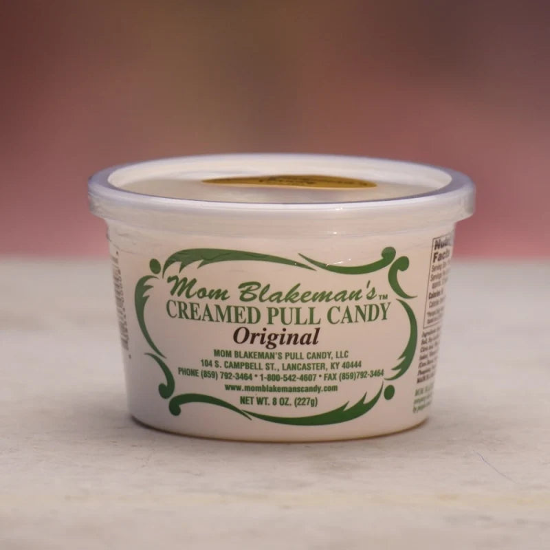 A 6 oz plastic tub of Mom Blakeman's Original Creamed Pull Candy, featuring a white container with a clear lid and green decorative text, set against a soft pink background.