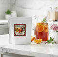 A white stand-up pouch of Gahma Apothecary Elderberry Lemon Cold Tea Blend sits on a marble countertop next to a glass pitcher filled with iced tea, orange slices, and mint, with loose oranges and a pink rose nearby.