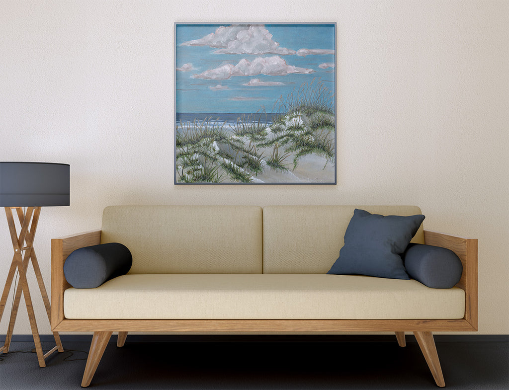 A framed square canvas print of a beach dune landscape with blue sky and clouds, displayed on a beige wall above a light wood sofa with dark blue pillows.