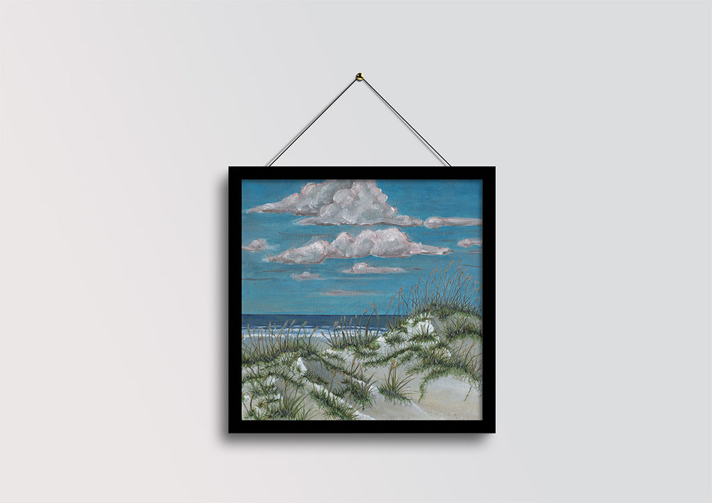 A framed square art print depicting a beach dune landscape with sand grasses, ocean, and blue sky with white clouds, hanging from a black string against a light gray wall.