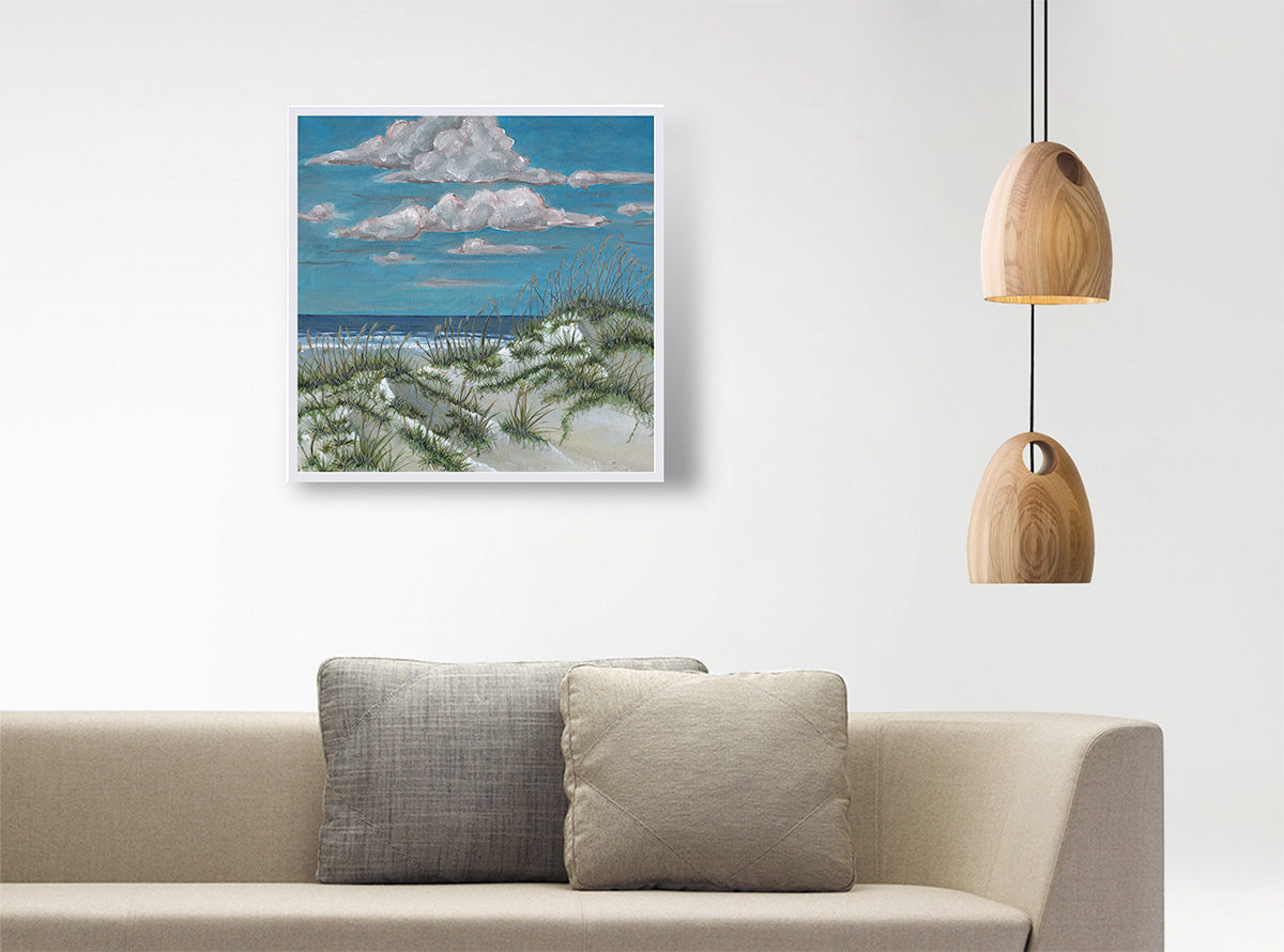 A square framed beach landscape painting hangs on a white wall above a beige sofa with two gray pillows. The painting depicts sand dunes, sea grass, ocean, and a blue sky with white clouds.