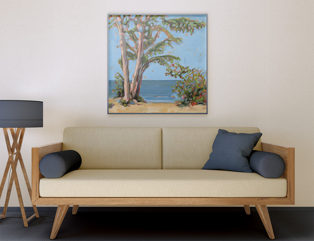 A framed painting of a beach scene with trees and blue water hangs on a beige wall above a light wood sofa with cream cushions and dark blue pillows.