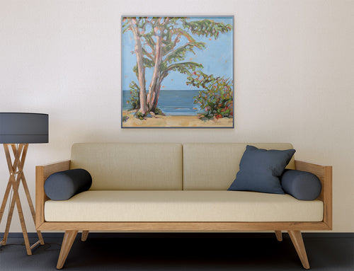 A framed painting of a beach scene with trees and blue water hangs on a beige wall above a light wood sofa with cream cushions and dark blue pillows.