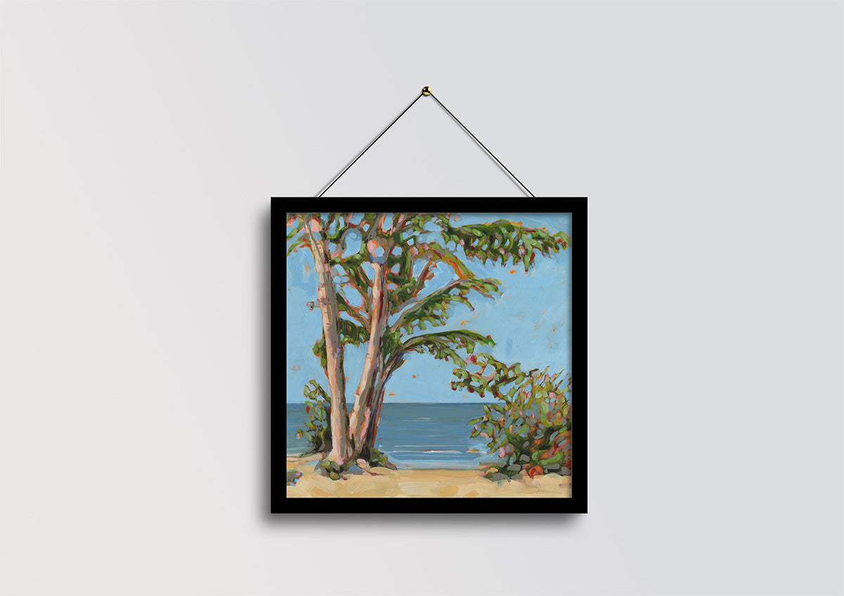 A framed landscape painting of coastal trees and shrubs by the sea, hanging on a light gray wall with a black frame and string.
