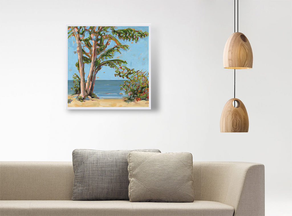 A framed square painting of a beach scene with trees and blue water, displayed on a white wall above a beige sofa with two gray pillows, next to two hanging wooden pendant lights.