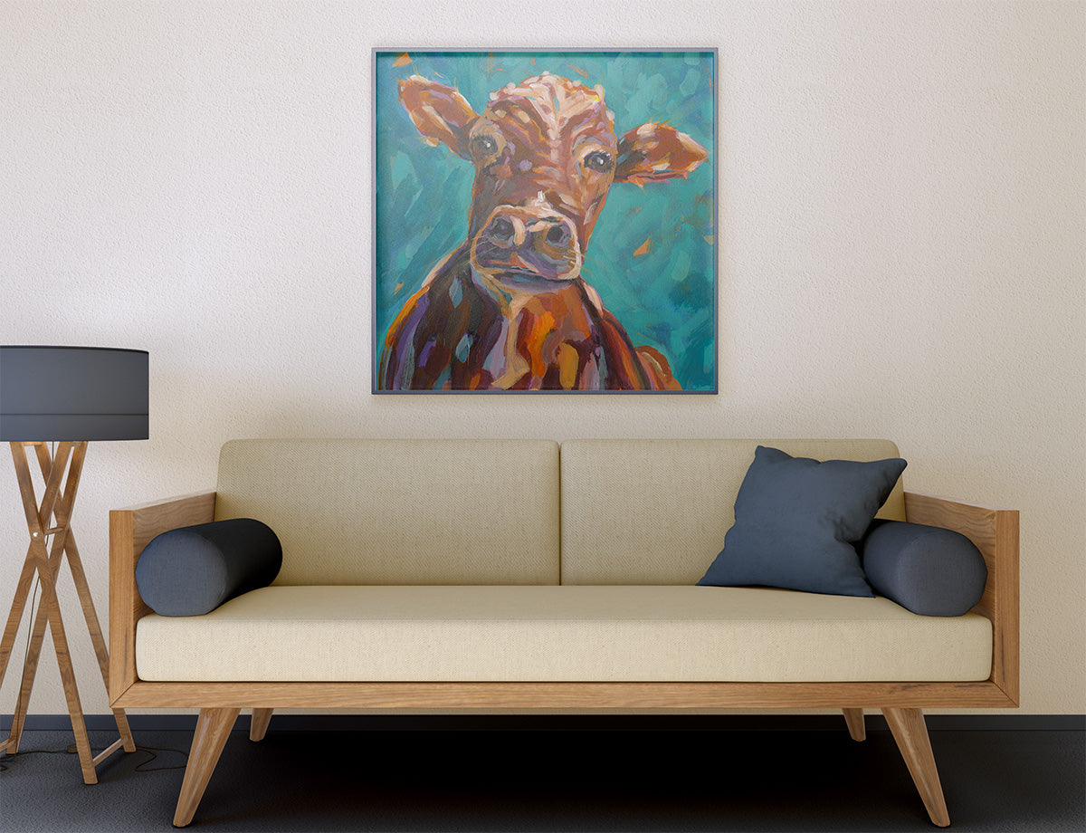 A framed square canvas painting of a brown cow on a teal background, displayed on a beige wall above a light wood sofa with dark blue pillows in a modern living room setting.