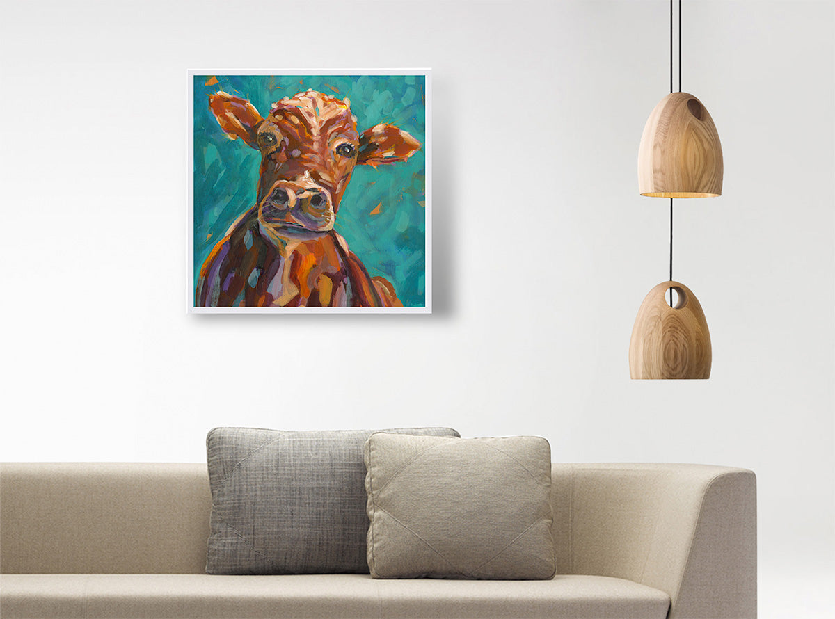 A square canvas art print featuring a colorful, textured oil painting of a brown cow on a teal background, styled on a white wall above a beige sofa with two pillows, next to a hanging wooden pendant light.