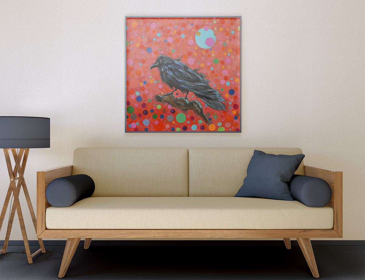 A framed square art print depicting a black raven perched on a branch against a vibrant red background with multicolored dots, hanging on a beige wall above a light wood sofa with dark blue pillows.