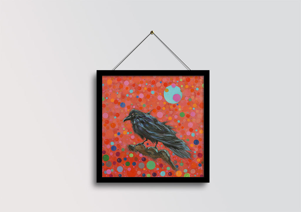 A framed square art print featuring a black raven perched on a branch against a vibrant red background filled with multicolored polka dots, hanging from a black wire on a light gray wall.