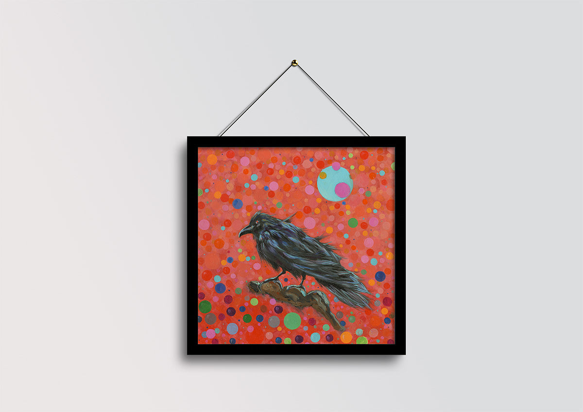 A framed square art print featuring a black raven perched on a branch against a vibrant red background filled with multicolored polka dots, hanging from a black wire on a light gray wall.