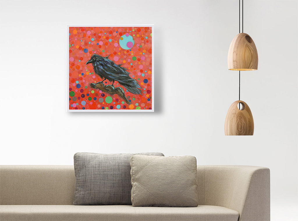 A framed square art print featuring a black raven on a vibrant red-orange background with colorful dots, displayed on a white wall above a beige sofa with two gray pillows, next to a hanging wooden pendant light.