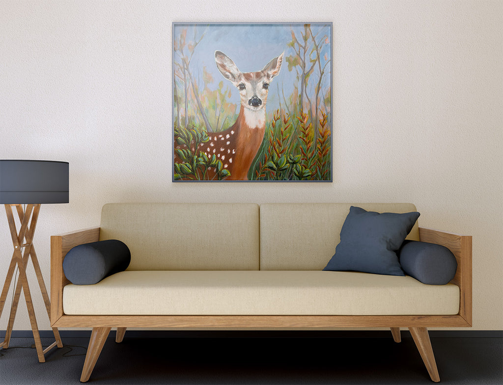 A framed painting of a deer in a forest hangs on a beige wall above a light wood sofa with dark blue pillows in a modern living room setting.