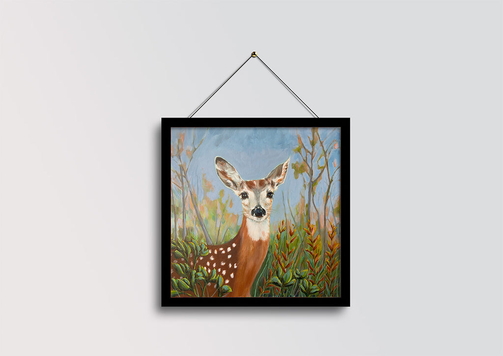 A framed art print featuring a painted deer in a grassy field, hanging from a black string against a light gray wall.