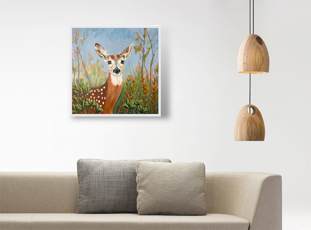 A framed art print of a deer in a field hanging on a white wall above a beige sofa with two gray pillows, next to a hanging wooden pendant light.