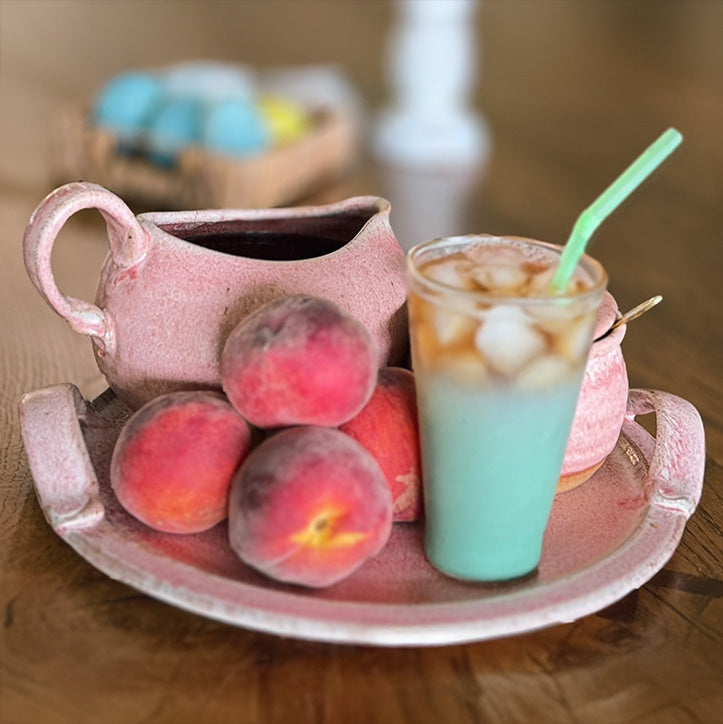 A pink ceramic pitcher and matching tray styled with fresh peaches and a layered blue-green iced drink with a green straw on a wooden table.