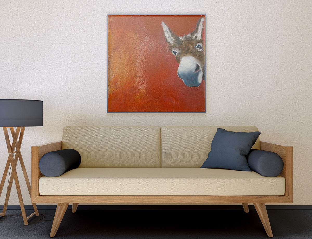 A framed square wall art print featuring a close-up donkey portrait on a warm orange-red background, displayed above a beige sofa in a modern living room setting.