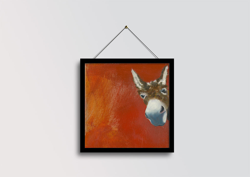 A framed square painting of a donkey's head against a textured orange-red background, hanging from a thin cord on a light gray wall.