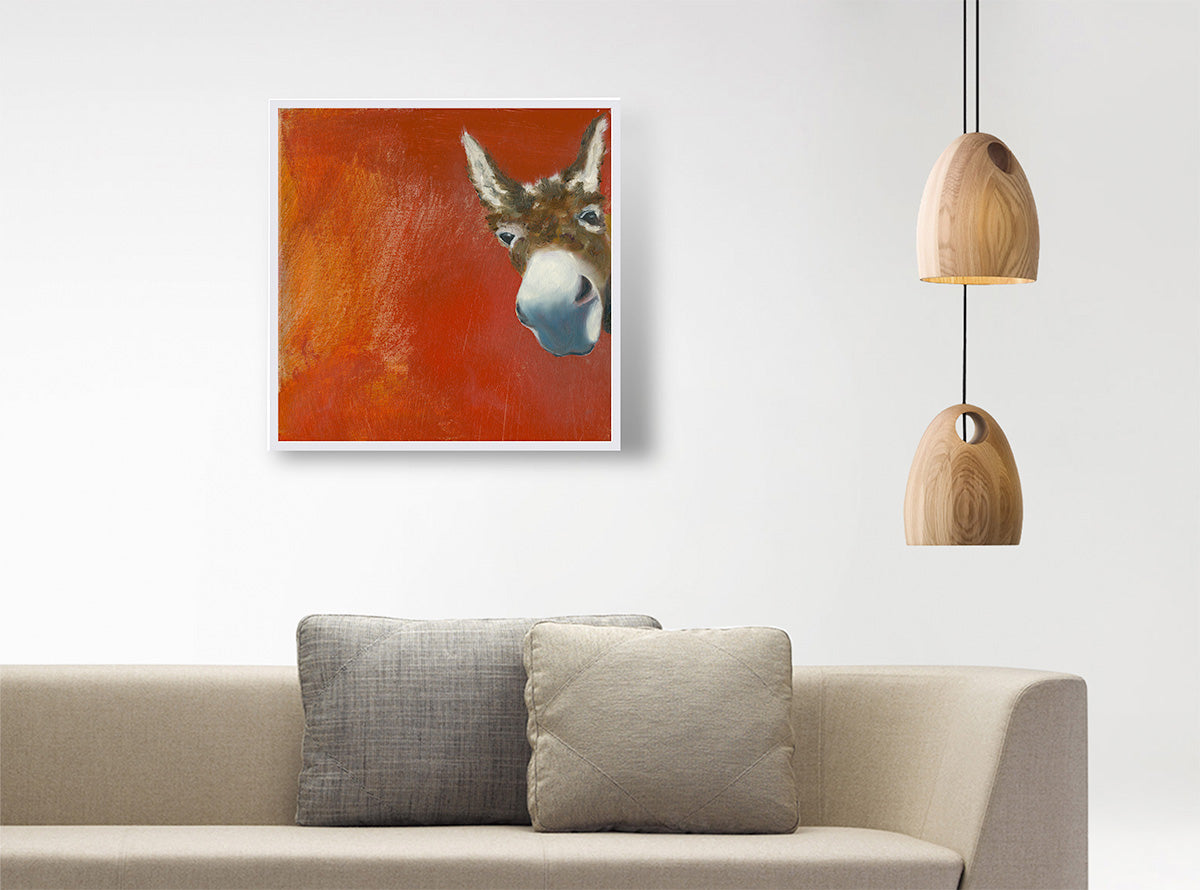 A square framed wall art print featuring a painted donkey head on a vivid orange-red background, displayed above a beige sofa with two pillows in a modern living room setting with hanging pendant lights.