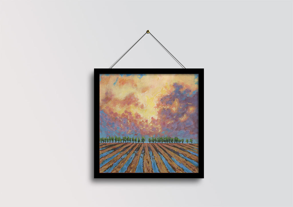 A framed landscape painting of a field under a dramatic sunset sky, hanging from a black string against a light gray wall.