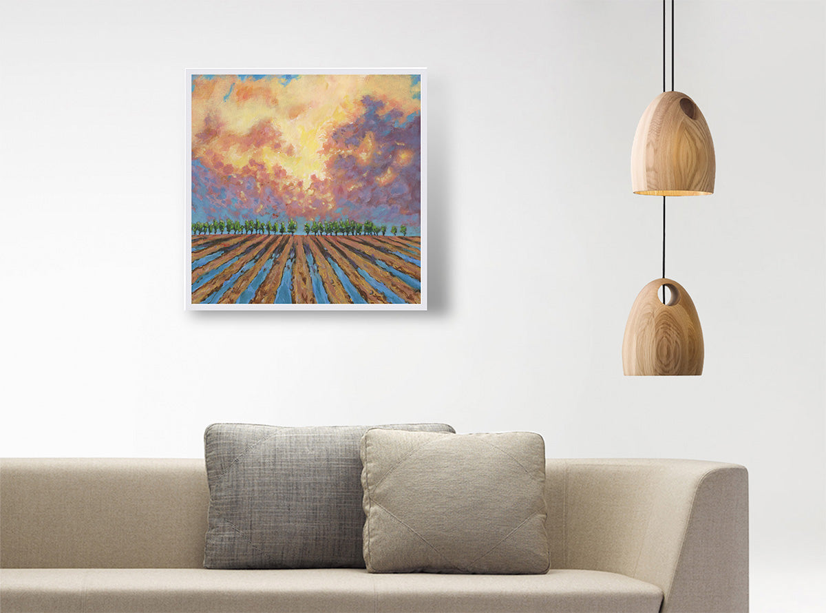 A square framed painting of a plowed field under a dramatic orange, yellow, and purple sky, displayed on a white wall above a beige sofa with two pillows, next to a hanging wooden pendant light.