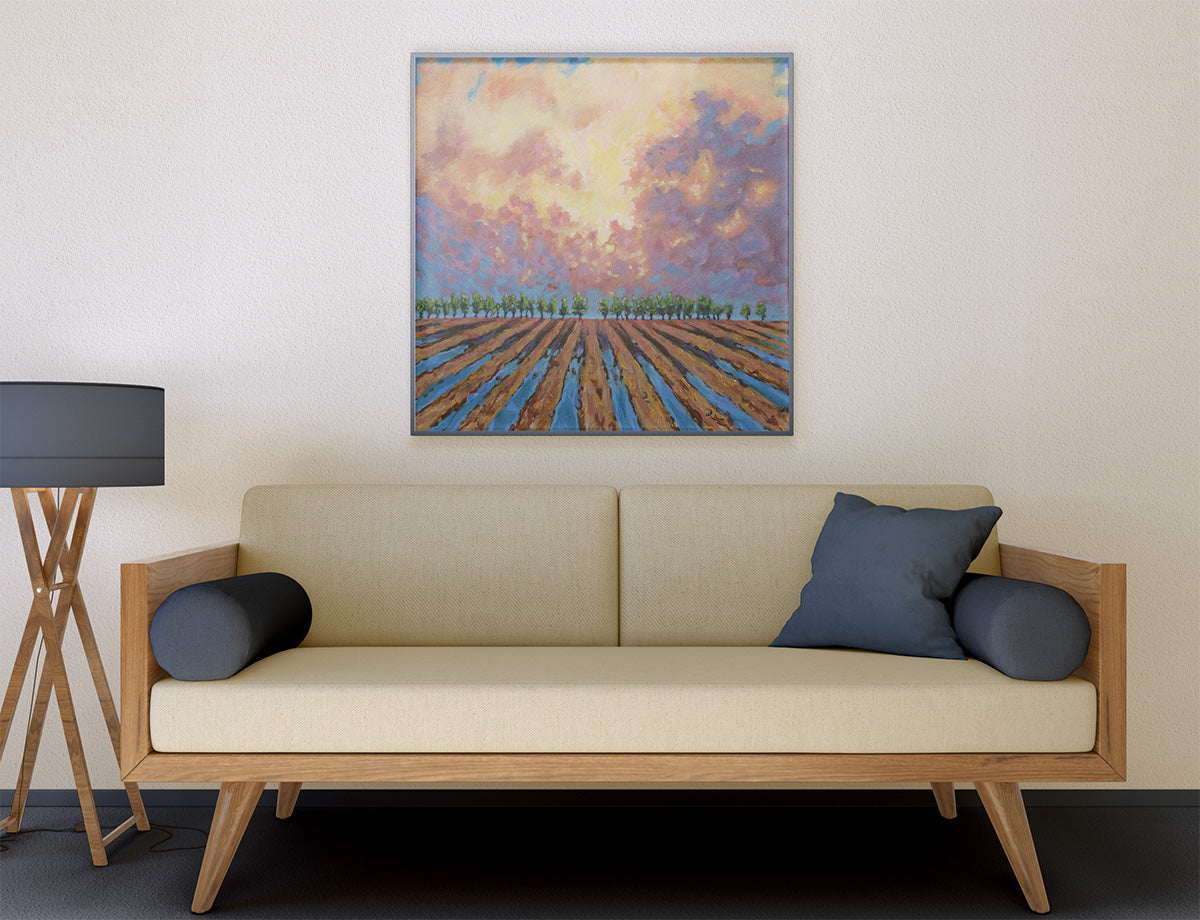 A framed landscape painting of a plowed field under a colorful sky, displayed on a beige wall above a light wood sofa with dark blue pillows in a modern living room setting.