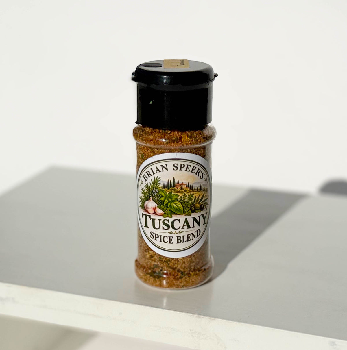 A clear plastic shaker bottle of Brian Speers Tuscany Spice Blend with a black lid, standing on a white surface against a white background.
