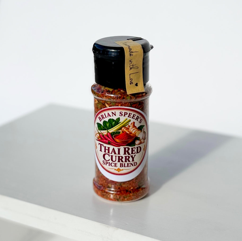 A bottle of Brian Speers Thai Red Curry Spice Blend on a white surface with a soft shadow. The clear bottle shows the red and orange spice mix inside, with a black cap and a yellow paper tag attached.