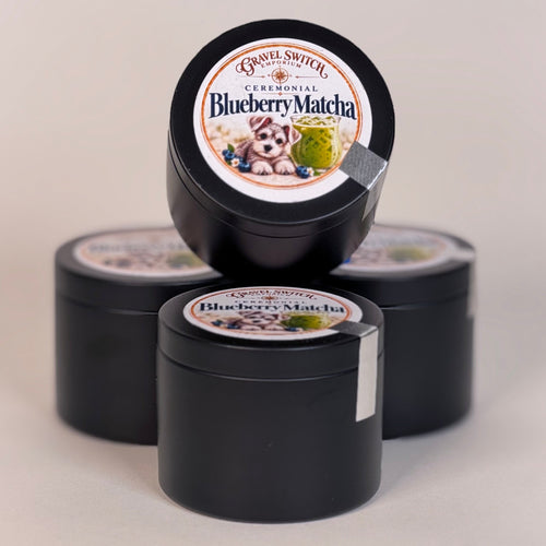 Three small round black tins of Genuine Snigle Ceremonial Blueberry Matcha, with one tin standing upright in front and the other two behind it, on a light beige background.
