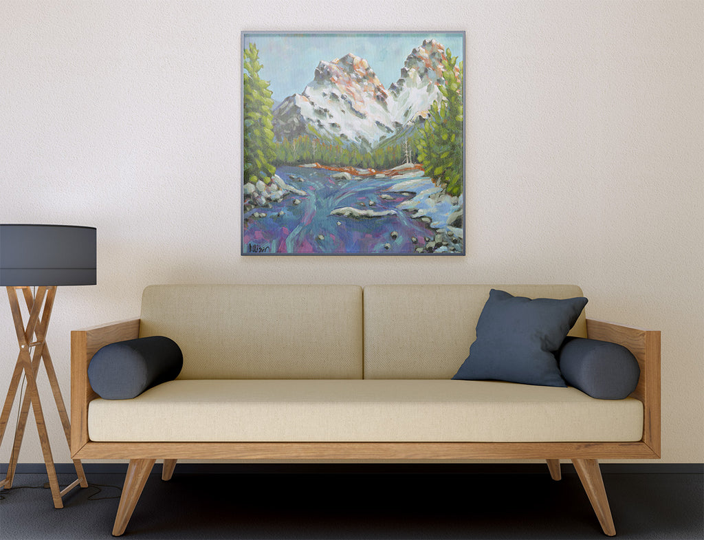 A framed landscape painting of a mountain river scene hangs on a beige wall above a light wood sofa with dark blue pillows in a modern living room setting.