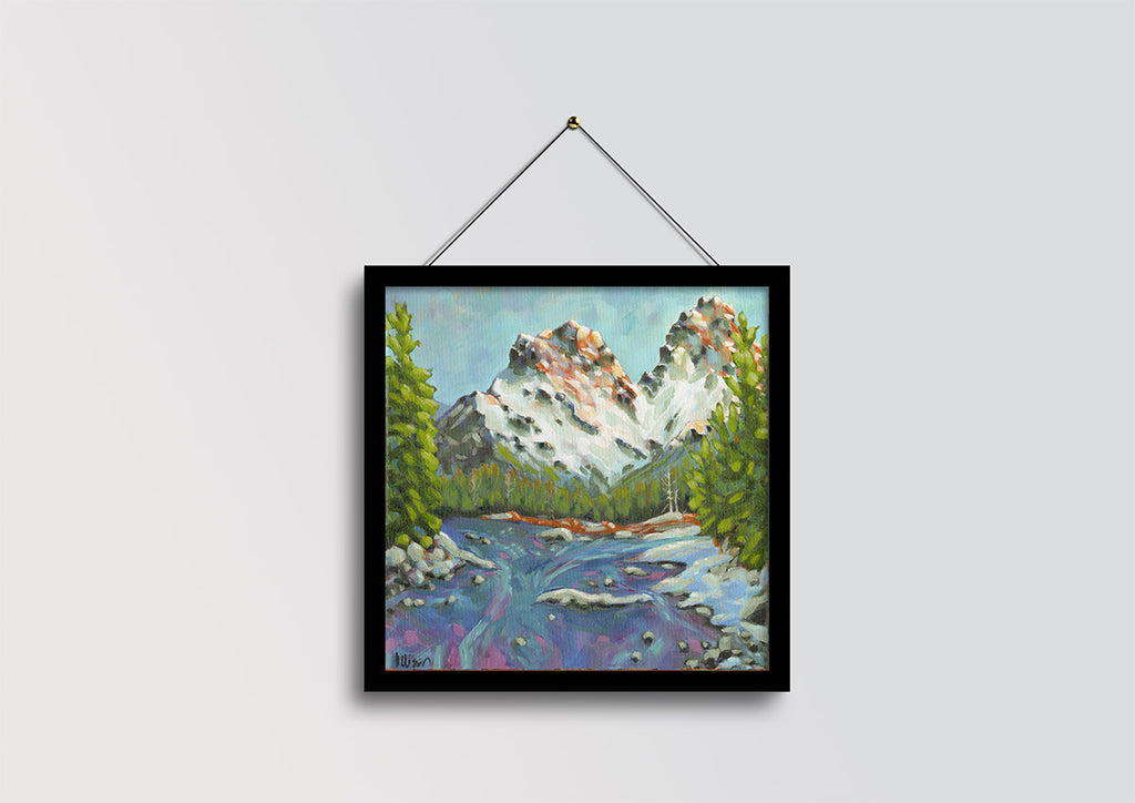 A framed landscape painting of a mountain scene with a river, hanging on a light gray wall by a black string.