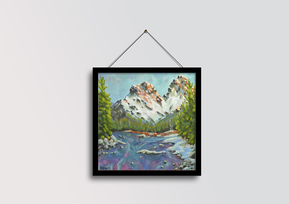 A framed landscape painting of a mountain scene with a river, hanging on a light gray wall by a black string.