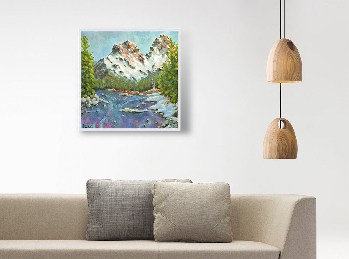 A square framed landscape painting of a mountain range with a river and trees, displayed on a white wall above a beige sofa with two gray pillows, with two wooden pendant lights hanging on the right.