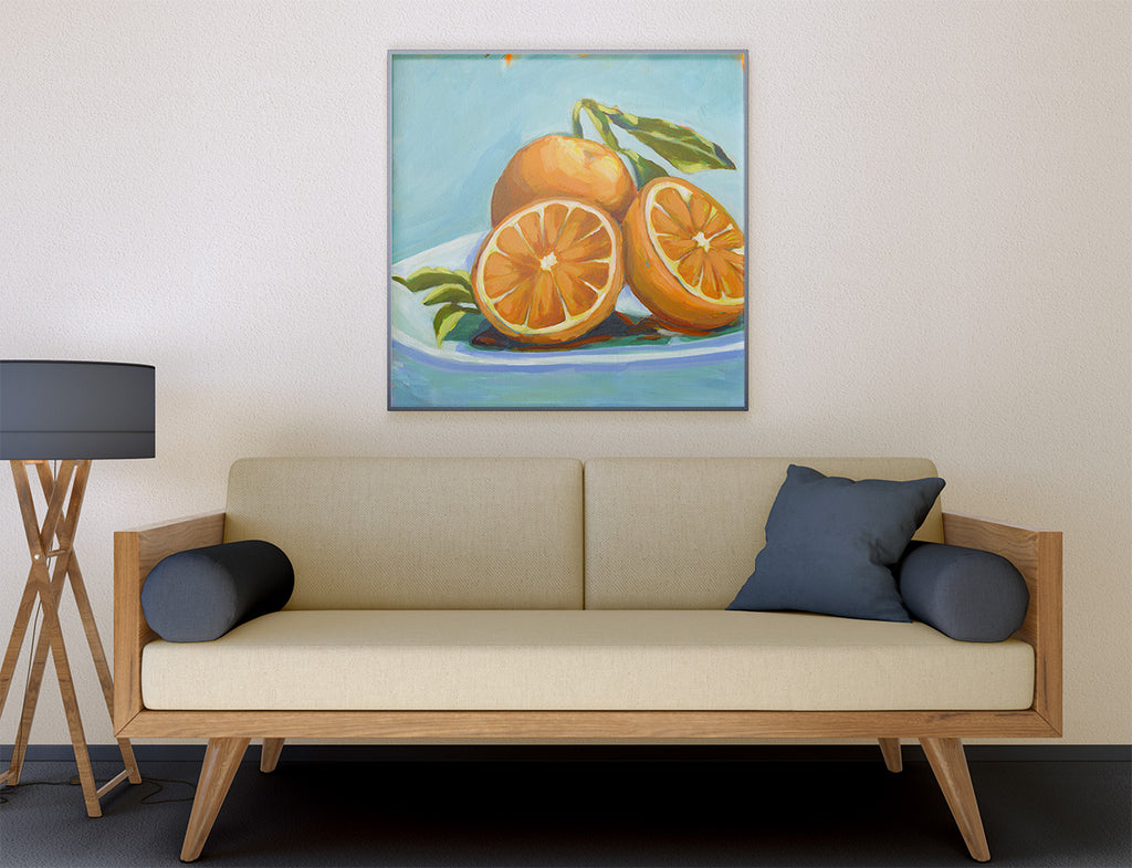 A framed still life painting of oranges on a plate, displayed on a beige wall above a light wood sofa with dark blue pillows in a modern living room setting.