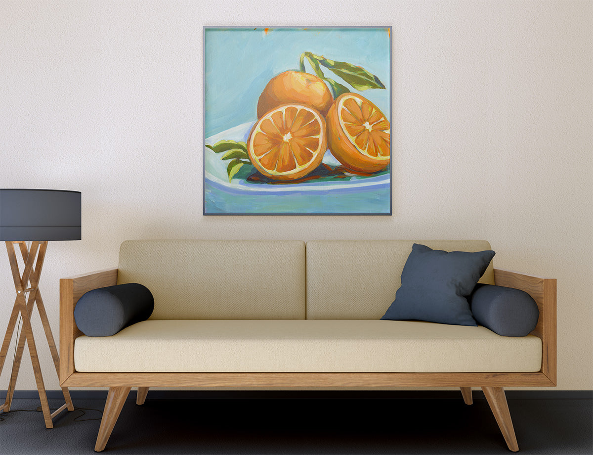 A framed still life painting of oranges on a plate, displayed on a beige wall above a light wood sofa with dark blue pillows in a modern living room setting.