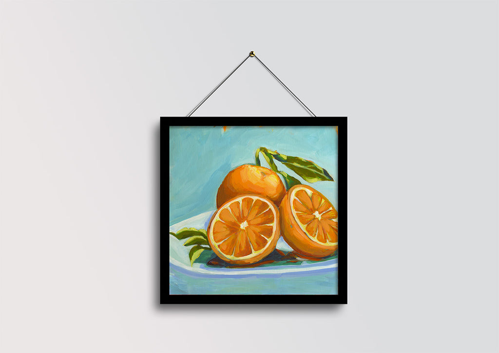A framed still life painting of oranges on a plate, hanging against a light gray wall with a black frame and string.