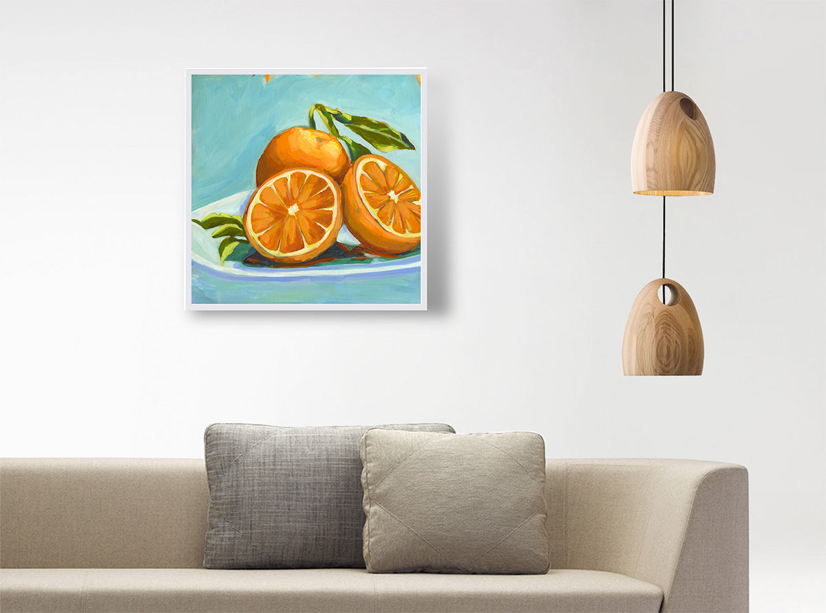 A square canvas painting of oranges on a plate, hanging on a white wall above a beige sofa with two gray pillows. Two wooden pendant lights hang on the right side of the wall.
