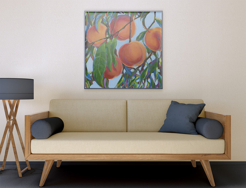 A framed square painting of peaches on branches hangs on a beige wall above a light wood sofa with cream cushions and dark blue pillows. A black floor lamp stands to the left.
