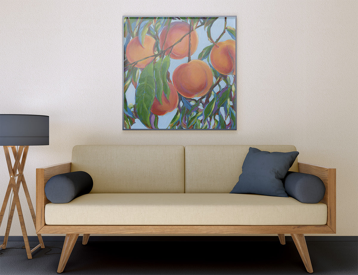 A framed square painting of peaches on branches hangs on a beige wall above a light wood sofa with cream cushions and dark blue pillows. A black floor lamp stands to the left.