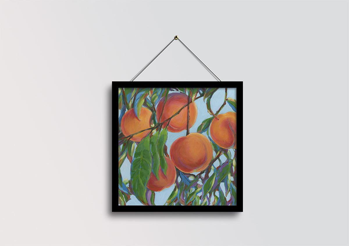 A framed painting of ripe oranges hanging from a tree branch with green leaves against a light blue sky, displayed on a light gray wall with a hanging string.