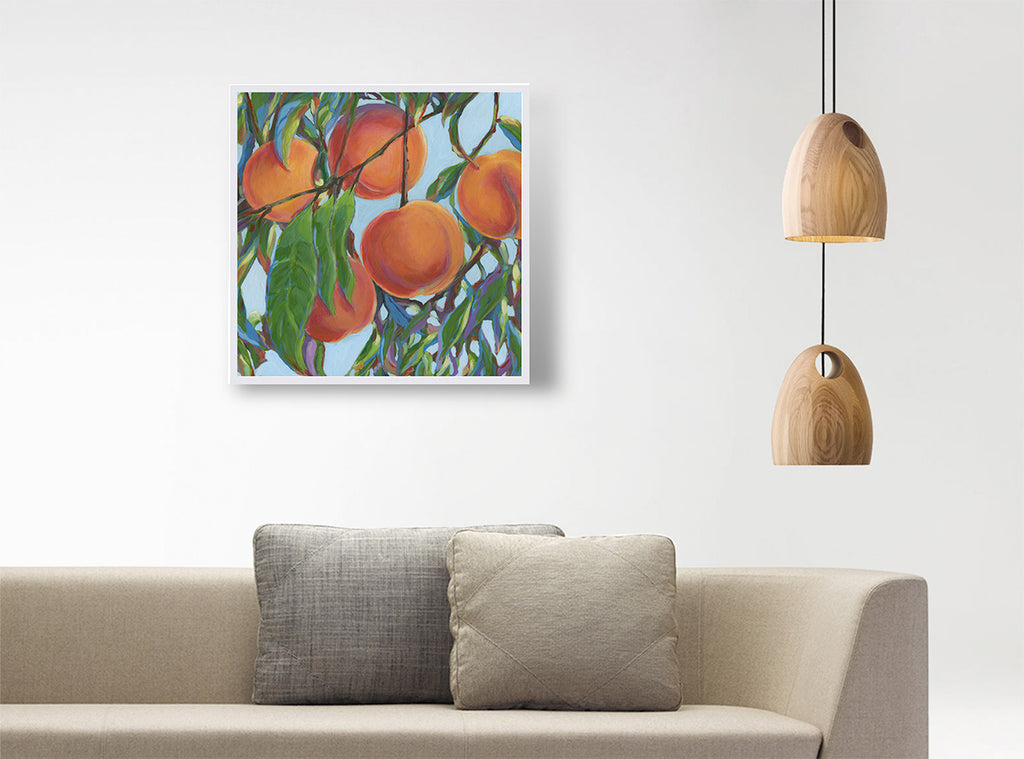 A square framed painting of peaches on a tree with green leaves and blue sky, displayed on a white wall above a beige sofa with two gray pillows, with two hanging wooden pendant lights on the right.