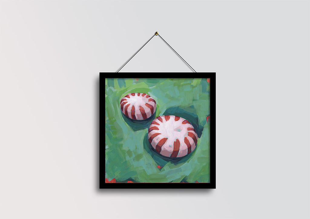 A framed square art print depicting two red-and-white striped peppermint candies on a textured green background, hanging from a black cord against a light gray wall.