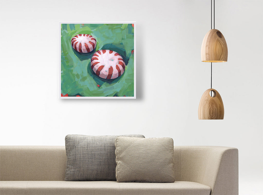 A square framed wall art print depicting two red-and-white striped candy-like motifs on a green background, displayed above a beige sofa with two gray pillows in a modern interior setting with hanging wooden pendant lights.