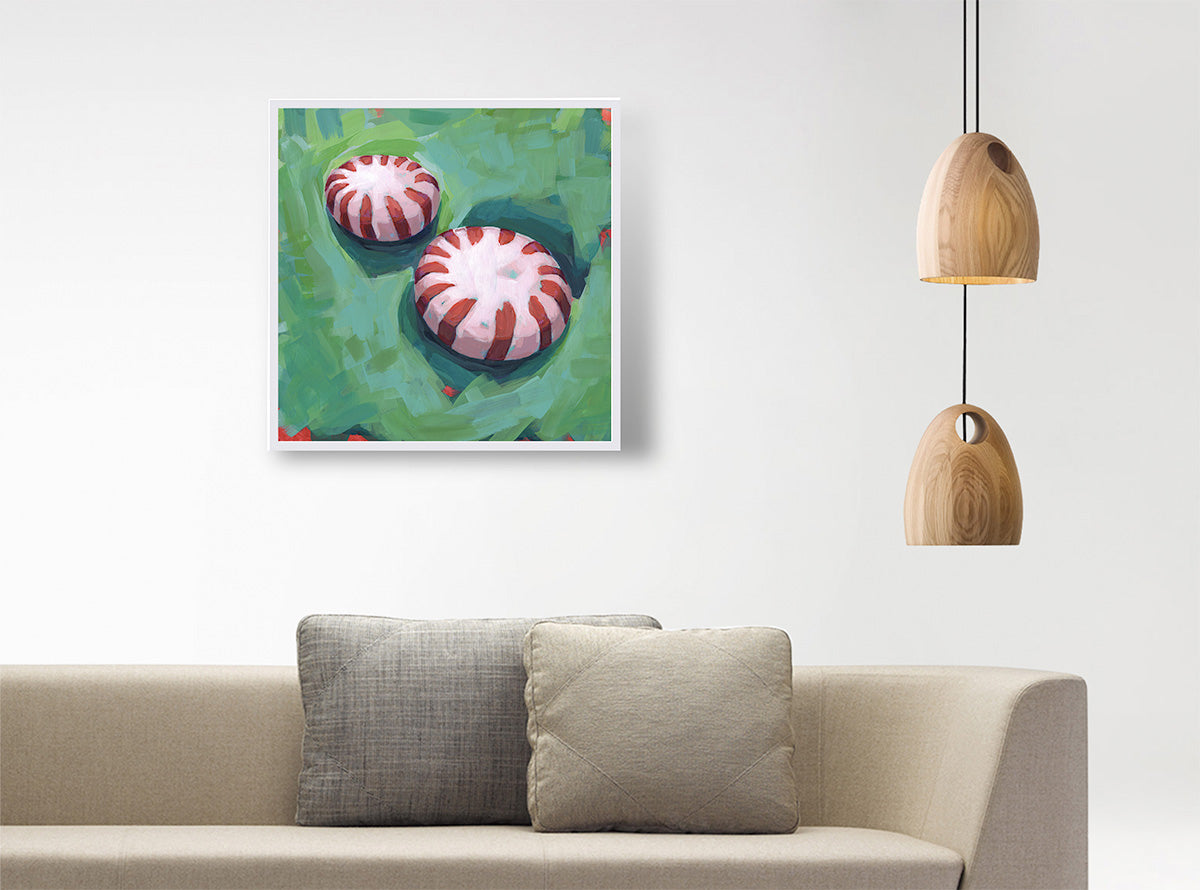 A square framed wall art print depicting two red-and-white striped candy-like motifs on a green background, displayed above a beige sofa with two gray pillows in a modern interior setting with hanging wooden pendant lights.