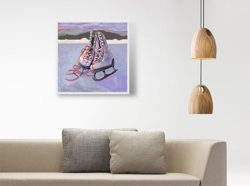 A framed art print depicting a pair of ice skates on an icy surface, hanging on a white wall above a beige sofa with two pillows, next to a hanging wooden pendant light.