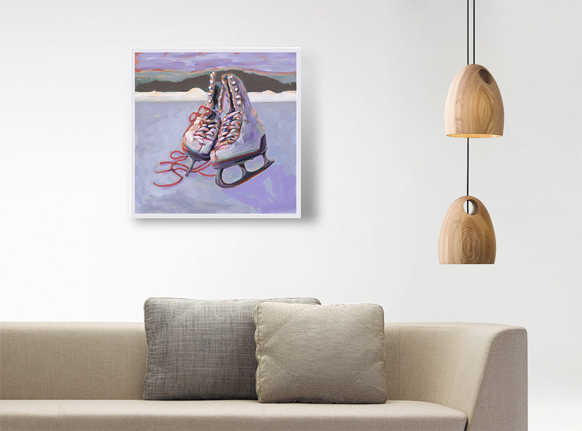 A framed art print depicting a pair of ice skates on an icy surface, hanging on a white wall above a beige sofa with two pillows, next to a hanging wooden pendant light.