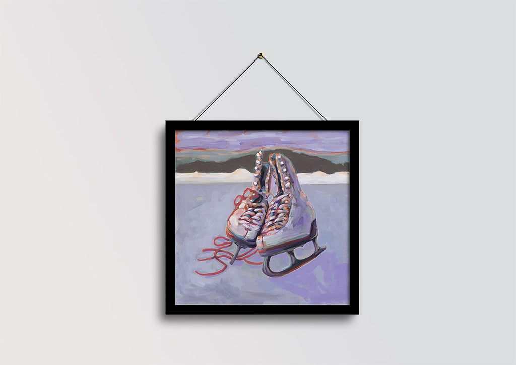 A framed art print depicting a pair of ice skates on a purple-toned rink, hanging from a black string against a light gray wall.
