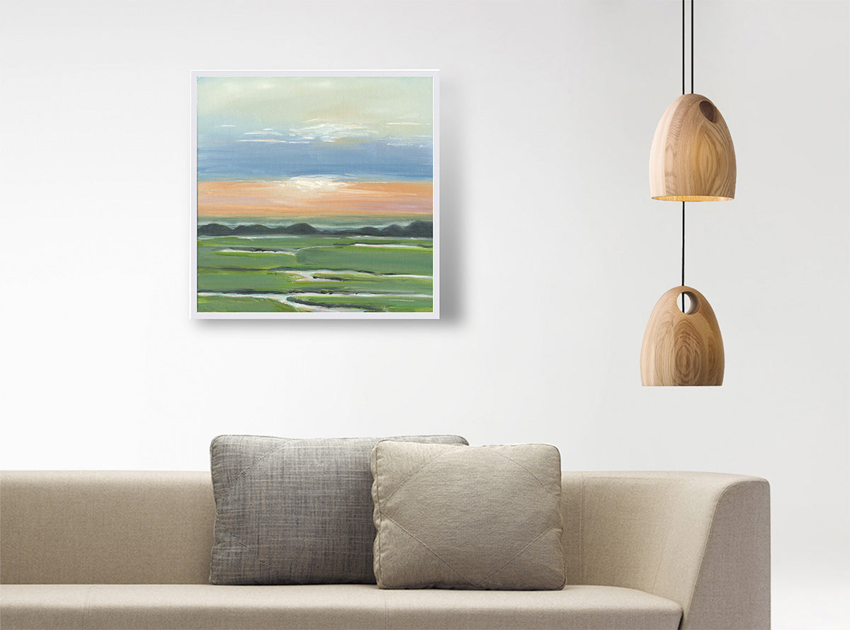 A framed landscape painting of green fields under a blue and orange sky, displayed on a white wall above a beige sofa with two gray pillows, next to two hanging wooden pendant lights.