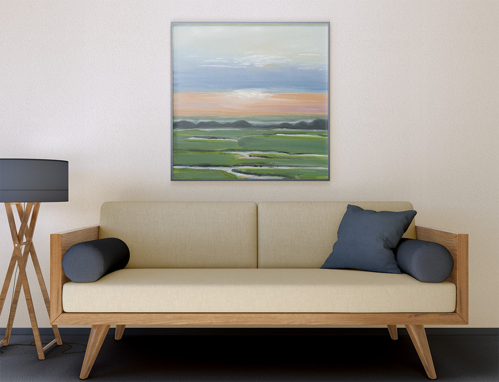 A framed landscape painting of a green field under a blue sky hangs on a beige wall above a mid-century modern wooden sofa with cream cushions and dark blue pillows.