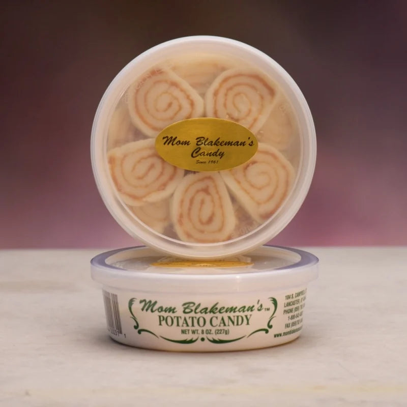 Two round plastic tubs of Mom Blakeman's Potato Candy stacked on a light surface against a dark purple gradient background. The top tub is open, showing spiral potato candy rolls inside, with a gold oval label.